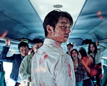 Gong Yoo spielt in Train to Busan den Fondsmanager Seok-woo. Dieser steht auf diesem Bild mit dem Rücken zum Betrachter und dreht seinen Kopf und den Oberkörper zur rechten Seite. Sein weißes Hemd weist mehrere Blutflecken auf. Im Hintergrund stehen noch mehrere Menschen.