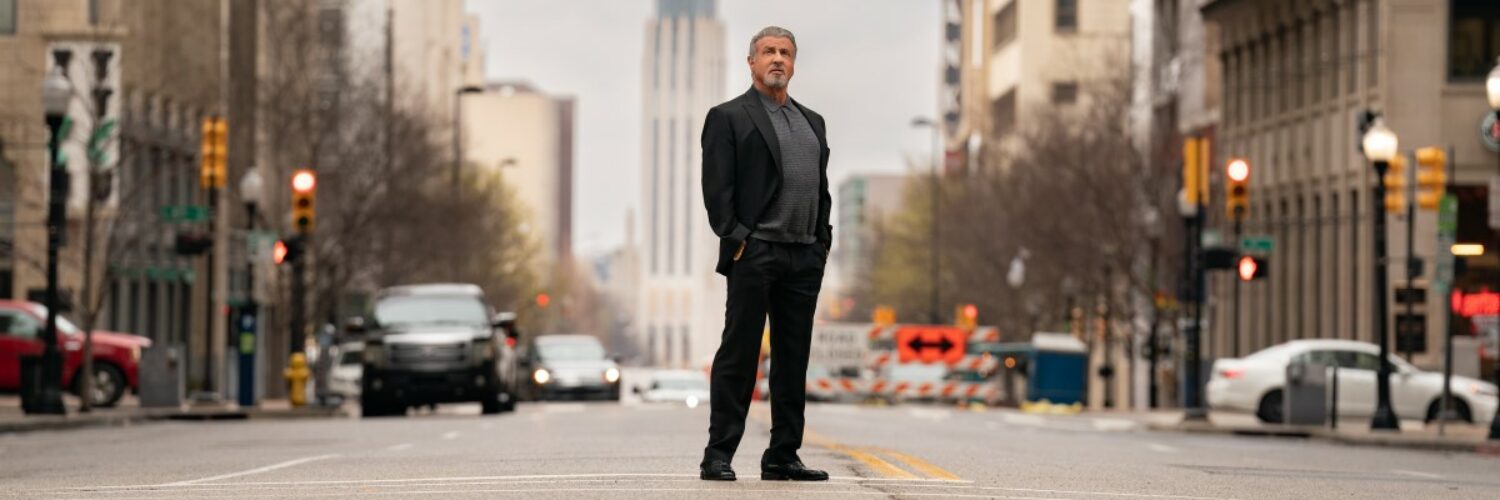 Sylvester Stallone im schwarzen Anzug in der Straßenmitte. Im Hintergrund Fahrzeuge, Hochhäuser