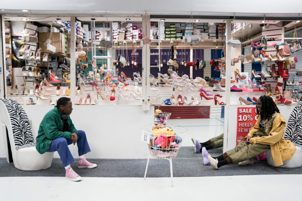 Dom und Yas sitzen auf überdimensionierten Schuhen in einer Mall