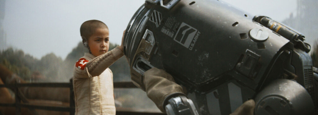 Ein kleines, glatzköpfiges Mädchen trägt ein beiges Oberteil. Es hält seine rechte Hand auf einen in Tarnfarbe gestrichenen, rundköpfigen Roboter, welcher sich zu ihr runterbeugt. Eine Szene aus dem Film The Creator.