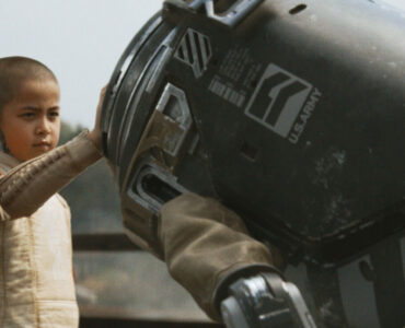 Ein kleines, glatzköpfiges Mädchen trägt ein beiges Oberteil. Es hält seine rechte Hand auf einen in Tarnfarbe gestrichenen, rundköpfigen Roboter, welcher sich zu ihr runterbeugt. Eine Szene aus dem Film The Creator.