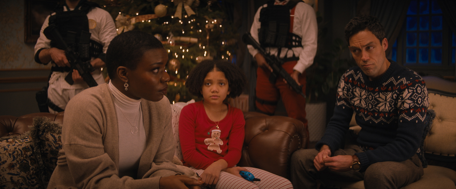 Alexis Louder als Linda Matthews sitzt ganz links auf dem Sofa und schaut besorgt zu ihrem Partner Jason Lightstone gespielt von Alex Hassell ganz rechts. Dieser schaut besorgt zurück, während Tochter Trudy Lightstone (Leah Brady)bin der Mitte, ebenfalls besorgt zu ihrer Mutter blickt. Hinter den dreien stehen zwei Leute mit Maschinengewehren vor dem Weihnachtsbaum im Hintergrund. 