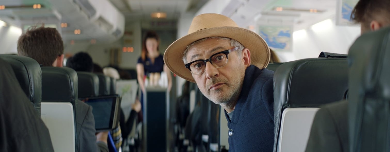 Elia Suleiman sitzt in einem Flugzeug, trägt eine schwarz gerahmte Brille und einen Strohhut und schaut in die Kamera. Im Hintergrund sieht man den Sitzgang des Flugzeugs, es kommt eine Stewardess mit einem Getränkewagen angelaufen.