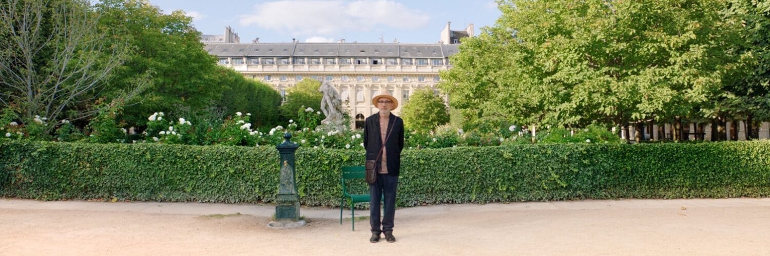 Elia Suleiman steht in einem dunklen Anzug und seinem Strohhut vor einer Grünanlage direkt vor einem alten, französischen Gebäude. Hinter ihm steht ein grüner Metallstuhl und ein grüner barocker Wasserspender. Er beobachtet etwas in der Ferne.