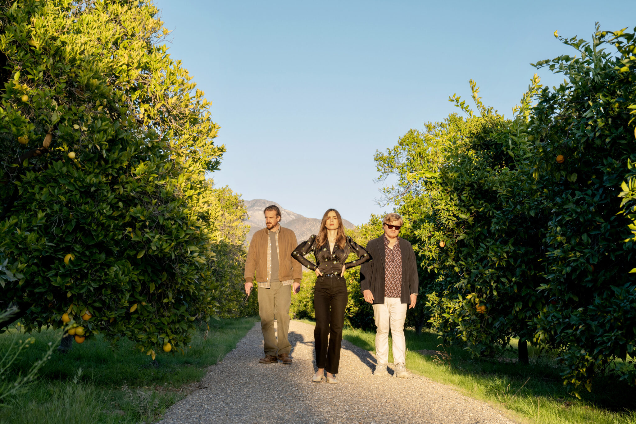 Drei Personen (Jason Segel, Lily Collins und Jesse Plemons) zwischen Orangenbäumen auf einem Kiesweg.