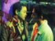 Ho Po-wing und Lai Yiu-fai eng zusammen hinter einer Glasfassade. Lai hat eine Zigarette zwischen den Lippen.