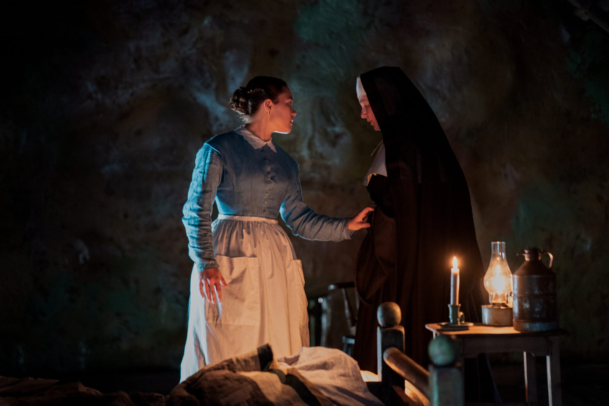 Florence Pugh in einem blauweißen Kleid neben einer Nonne in Schwarz in einer dunklen Kammer. Das Wunder
