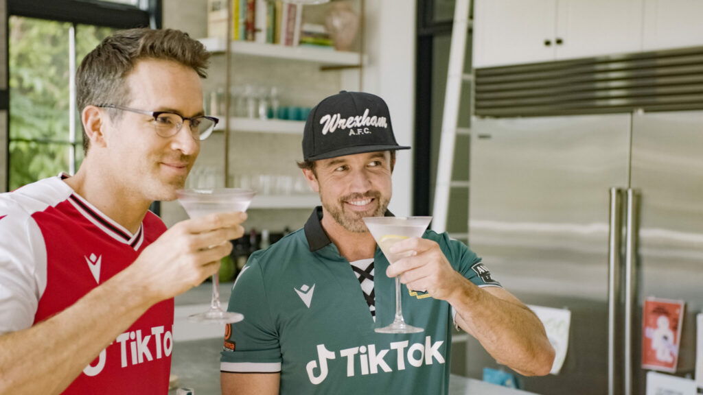 Ryan Reynolds und Rob McElhenney mit Trikots des AFC Wrexham und Cocktailgläsern in der Hand