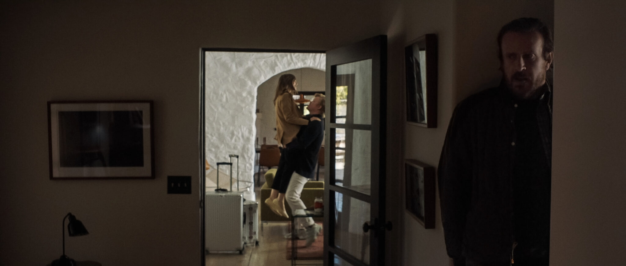 Im Vordergrund versteckt sich Jason Segel in einer Nische während hinten im Bild ein Pärchen im Eingangsbereich des Hauses steht. Die beiden haben Koffer neben sich stehen und der Mann (Jesse Plemons) hebt die Frau (Lily Collins) in die Höhe. Windfall