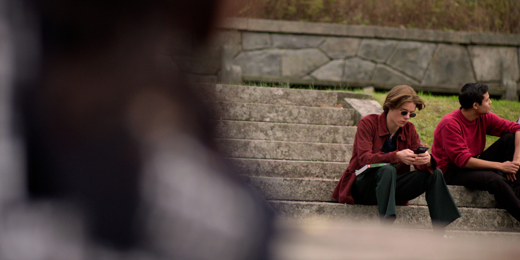 Ein junger Mann mit rotem Hemd und Sonnenbrille sitzt auf einer Treppe und tippt etwas in sein Smartphone ein.