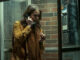Melody (Dina Shihabi) telefoniert in einer Telefonzelle vor dem Visser-Gebäude und schaut besorgt aus in Archive 81