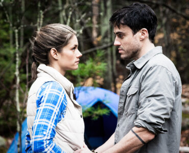 Die Hauptfiguren Jenn und Alex stehen vor ihrem Zelt im Wald und schauen sich an.
