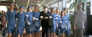 Leonardo DiCaprio läuft als Frank Abagnale im Piloten-Outfit mit acht Stewardessen in blauem Dress einen Gang im Flughafen entlang.