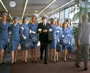 Leonardo DiCaprio läuft als Frank Abagnale im Piloten-Outfit mit acht Stewardessen in blauem Dress einen Gang im Flughafen entlang.