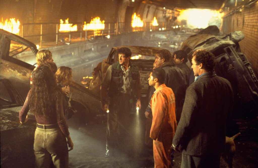 Stallone vor einer Gruppe Menschen im Tunnel. Im Hintergrund Autowracks und Brände.