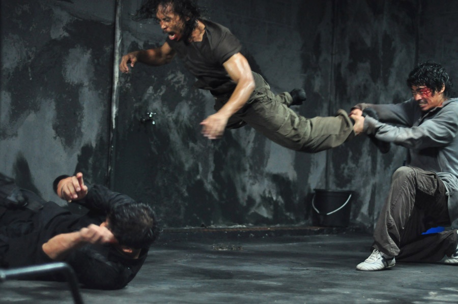 Andi (Donny Alamsyah) schleudert Gegner Mad Dog (Yayan Ruhian) an seinen Beinen herum, Protagonist Rama (Iko Uwais) windet sich schon auf dem grauen Betonboden - Die 15 coolsten Martial Arts Filme.