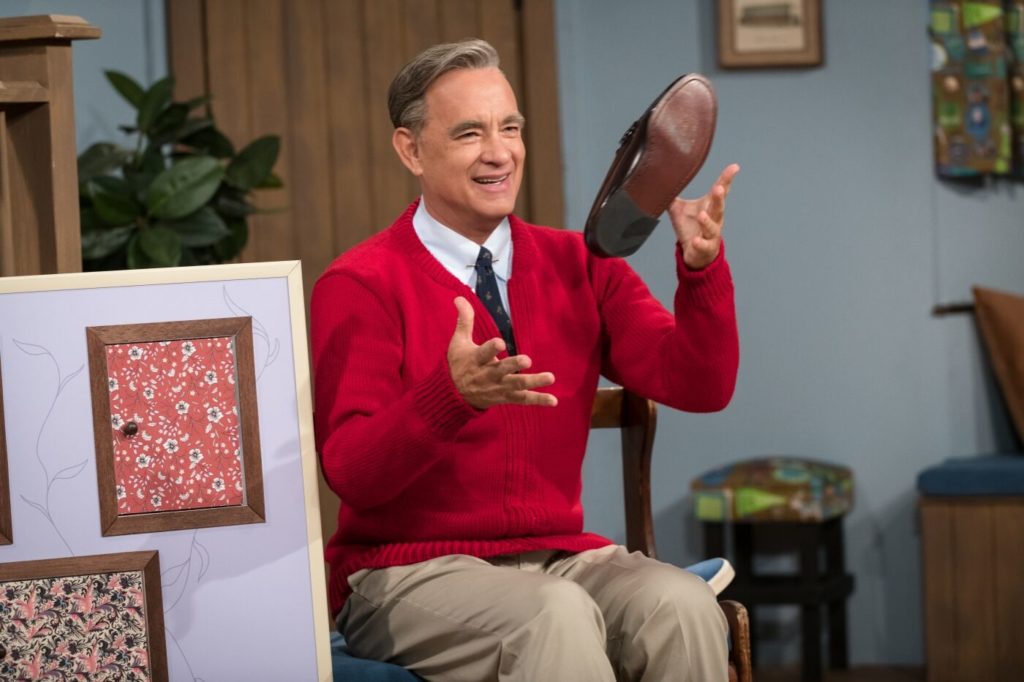 Tom Hanks als Fred Rogers, er jongliert mit einem Schuh und lacht dabei herzlich, er trägt einen roten Strickpullover und sitzt am Set der Serie in Der wunderbare Mr. Rogers