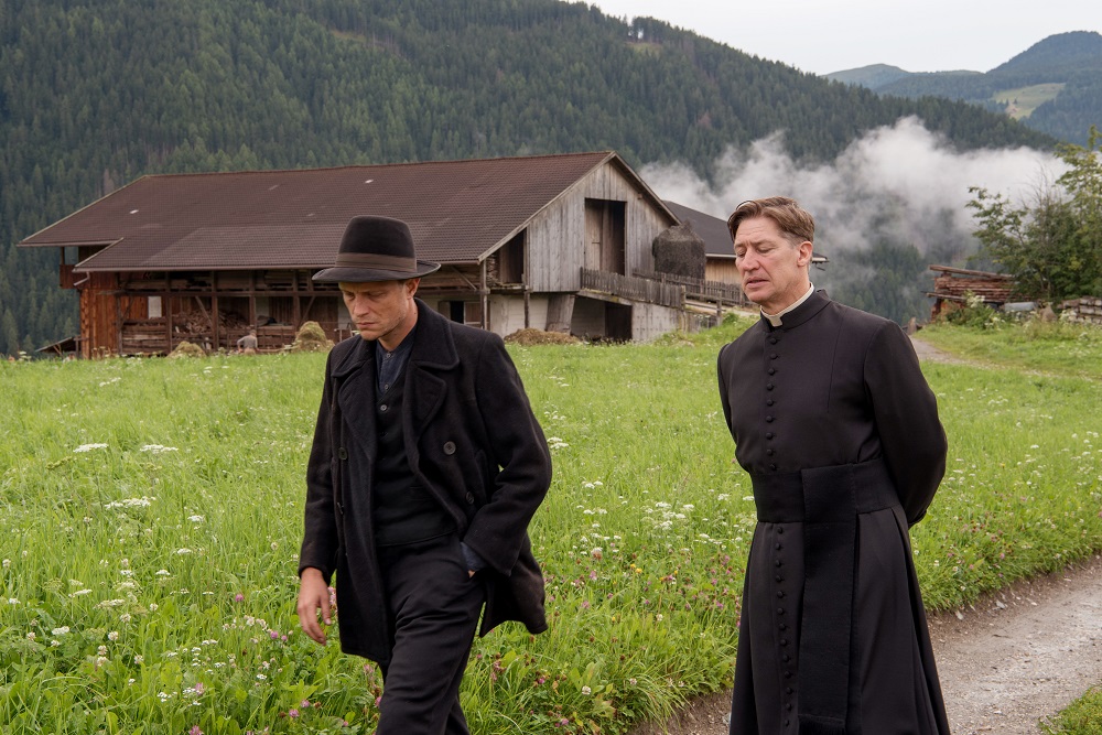 Franz Jägerstätter (August Diehl) und Pfarrer Ferdinand Fürthauer (Tobias Moretti) gehen nebeneinander her, während im Hintergrund eine grüne Wiese, eine alte Hütte und die österreichische Berglandschaft zu sehen ist.