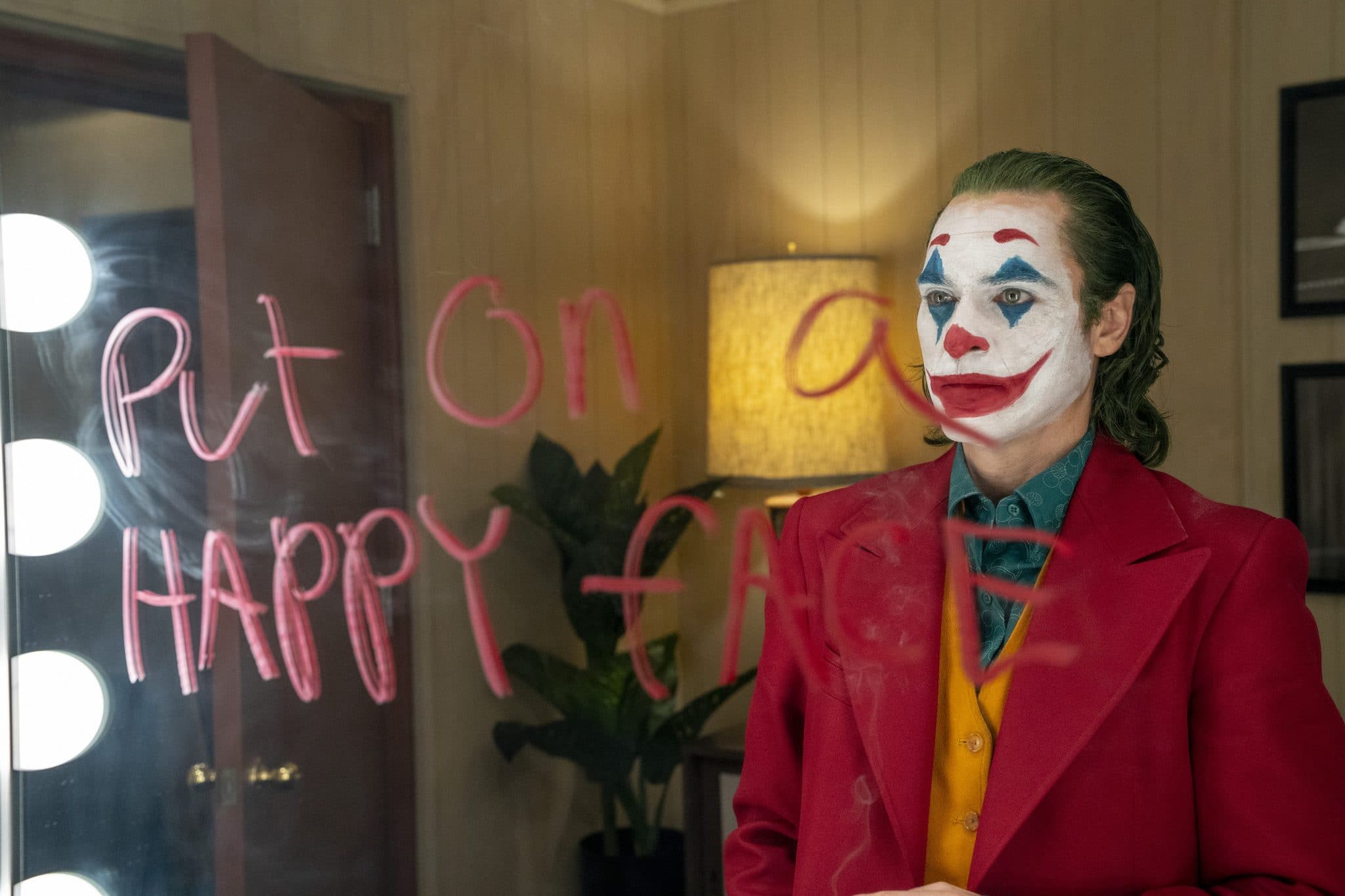 Arthur Fleck (Joaquin Phoenix) blickt im Joker-Kostüm in den Spiegel, auf dem in roter Farbe "Put on a happy face" geschrieben steht.