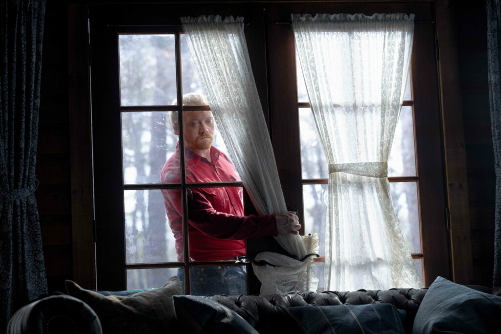 Redmond (Rupert Grint) trägt ein rotes Hemd und hat gerade ein Fenster der Hütte eingeschlagen. In Knock at the Cabin.