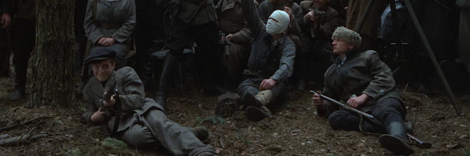 Dutzende Partisanen posieren für ein Foto. Einer trägt einen Verband um den Kopf und hält eine Handgranate hoch, während Fljora mit einem Gewehr auf dem Boden liegt.