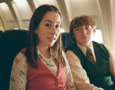 Alana und Gary sitzen nebeneinander im Flugzeug. Gary schaut Alana verliebt von der Seite an. Alana blickt den Gang entlang Richtung Kamera.
