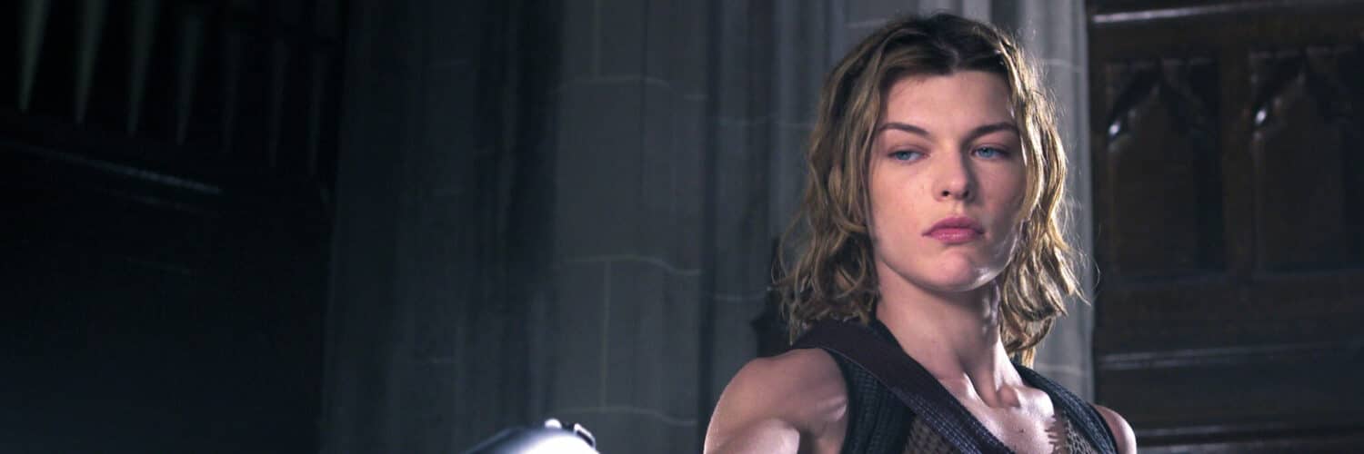Alice, gespielt von Milla Jovovich, steht in einem braunen Top und mit mehreren Waffen ausgestattet und richtet ihre Pistole auf einen Gegner am Boden
