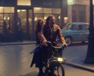 Victor (Daniel Auteuil) und Margot (Doria Tillier) fahren lachend mit einem Moped über die Straße, im Hintergrund ist die Kulisse des Cafés zu sehen.