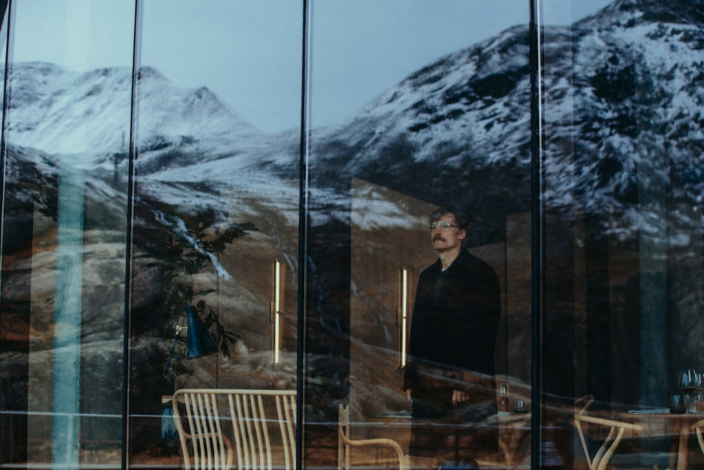 Max (Nikolaj Coster-Waldau) steht im Hotel vor einer Fensterwand und schaut hinaus auf die schneebedeckten Berge.