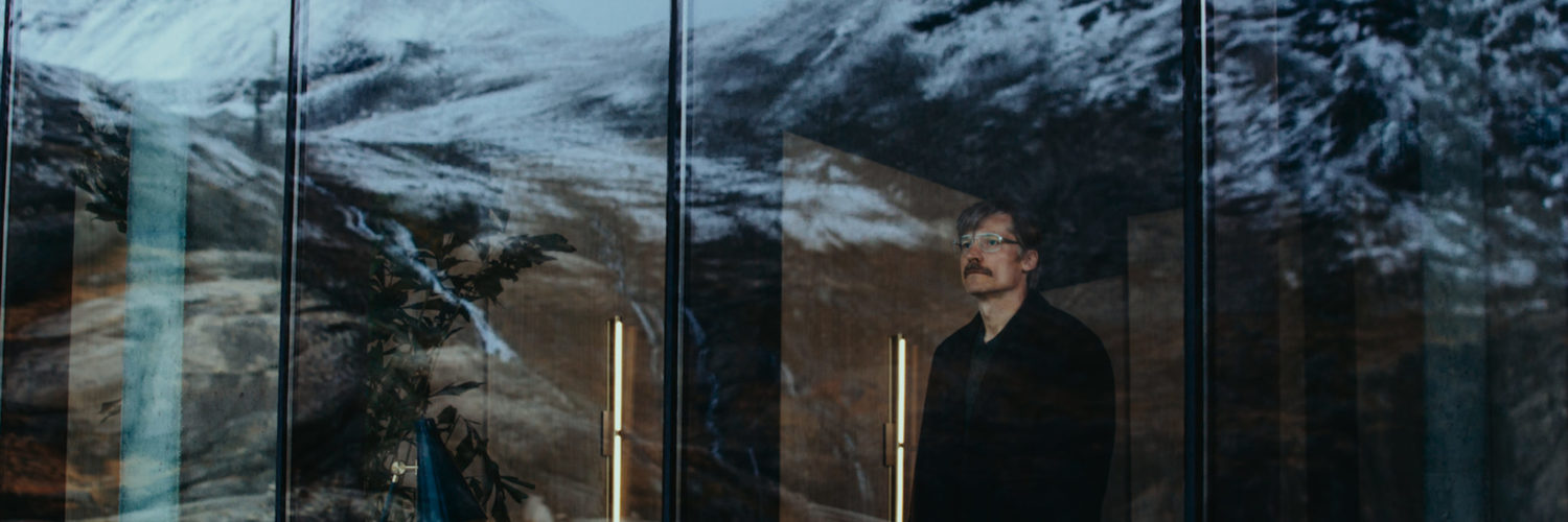 Max (Nikolaj Coster-Waldau) steht im Hotel vor einer Fensterwand und schaut hinaus auf die schneebedeckten Berge.