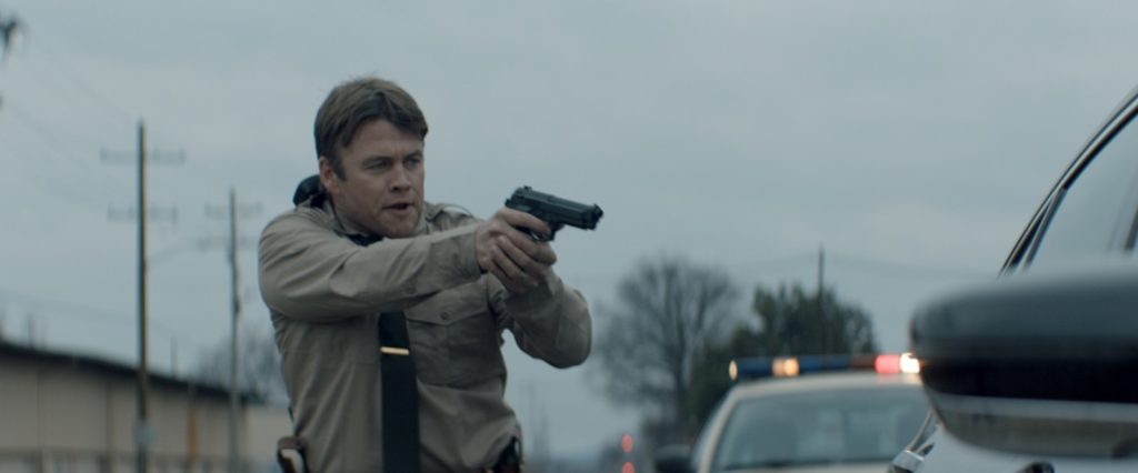 Luke Hemsworth als Cop steht in Tag der Vergeltung - Ein Vater sieht rot mit gezückter Waffe vor einem Auto.