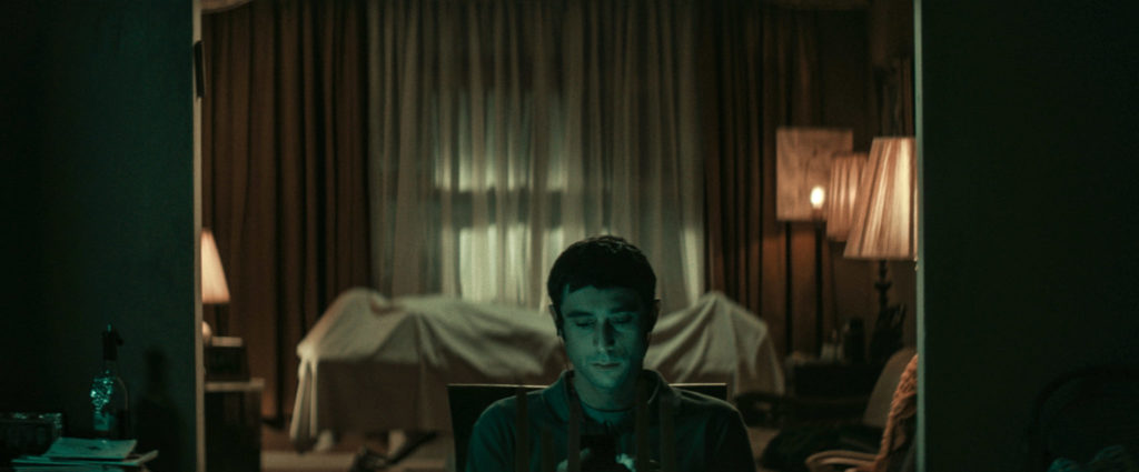Yakov (Dave Davis) starrt in THE VIGIL ausdruckslos in sein Handy, während hinter ihm die Leiche aufgebahrt ist.