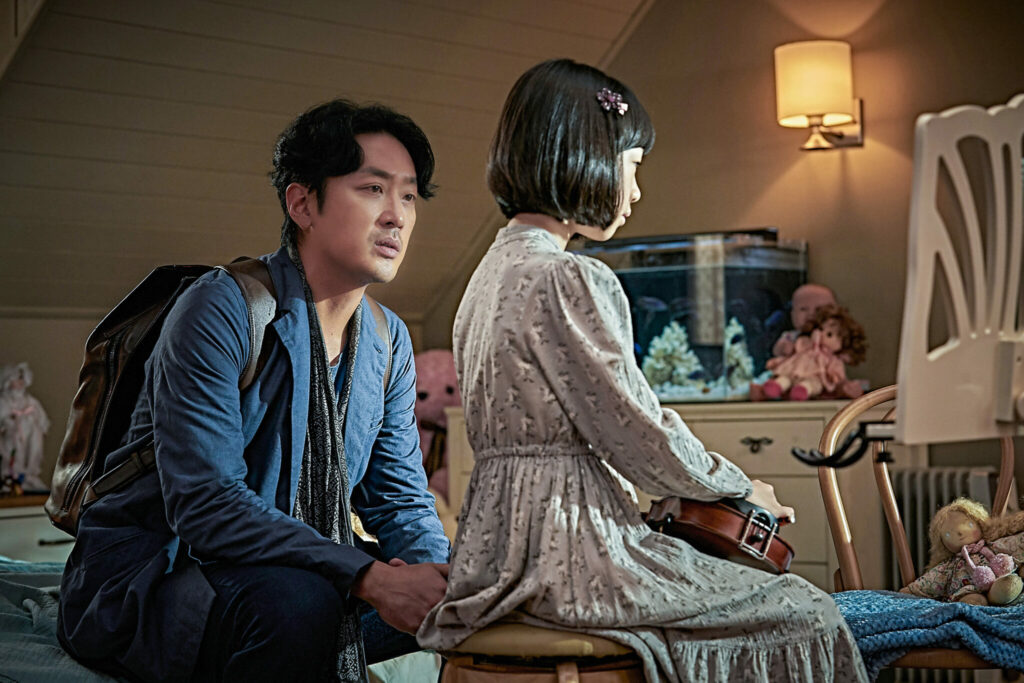 Sang-won sitzt auf dem Bett neben seinem Kind Ina, die eine Geige festhält in The Closet