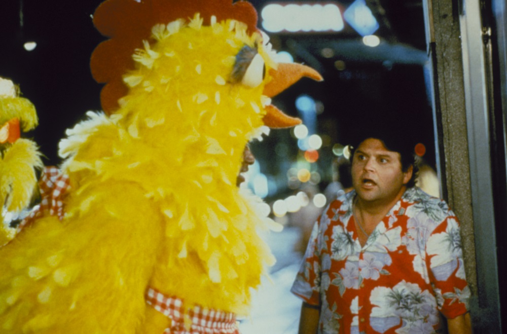 Links im Bild ist ein als Huhn verkleideter Mann, auf den Albert (Stephen Furst) rechts im Bild mit erschrockener Miene blickt.