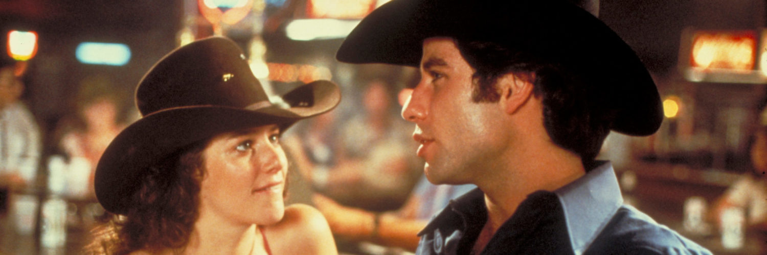 Debra Winger als Sissy blickt John Travolta als Bud Davis in "Urban Cowboy" leidenschaftlich an, während sich dieser an einem Bartresen mit ihr unterhält.